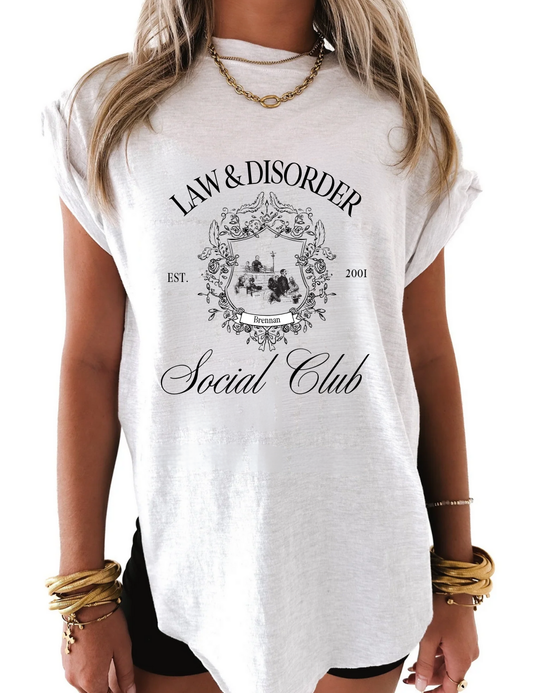Flat lay of white graphic tee that says “Law and Disorder” in bold black letters, folded on neutral background with chain-link accessories, Gen Z fashion.